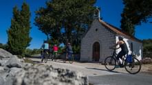 Gruppe von Radfahrern fährt an einer kleinen weißen Steinkapelle mit rotem Ziegeldach auf der Insel Šolta vorbei. Zypressen und blauer Himmel im Hintergrund.
