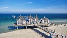 White pier building with ornate architecture extending into the Baltic Sea at Sellin, Rügen. Sandy beach and blue sky frame the historic structure.