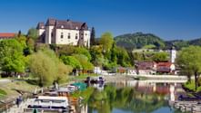 Grein Hafen mit Booten an der Donau. Greinburg thront prominent auf einem Hügel über der Stadt mit bewaldeten Bergen im Hintergrund.