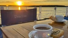 Coffee and pastries on wooden deck table with Princess chair at sunset over calm water. Golden sky reflects on the sea.