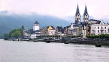 Bopparder Rheinufer mit historischen Gebäuden und zwei Kirchtürmen. Neblige Hügel im Hintergrund sichtbar.