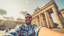 Lächelnder Mann mit Sonnenbrille macht Selfie vor dem Brandenburger Tor in Berlin. Das neoklassizistische Monument mit Säulen ist hinter ihm sichtbar.