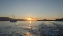 Sonnenuntergang über ruhigem Meer in Süddalmatien mit Segelboot und Inseln als Silhouetten vor orangefarbenem Himmel. Bootsspur im Vordergrund.