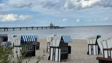 Striped wicker beach chairs on sandy beach in Zinnowitz with historic pier extending into the Baltic Sea under cloudy sky.