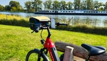 Red rental bicycle parked on wooden bench by river with cargo ship passing in background. Green grass and trees line the waterway.