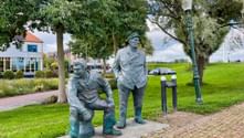 Zwei Bronzestatuen in einem Park in Medemblik: ein sitzender Mann und ein stehender Kapitän. Grüne Wiese und Bäume im Hintergrund mit Gebäuden.