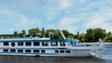 Weißes Flusskreuzfahrtschiff namens Switzerland fährt auf einem Fluss mit grünen Bäumen und blauem Himmel im Hintergrund. Das Schiff hat zwei Decks mit Fenstern.