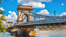 Chain Bridge spanning the Danube River in Budapest with stone pillars and suspension structure under a blue sky with white clouds.