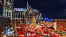 Beleuchteter Weihnachtsmarkt mit rot überdachten Ständen vor dem Kölner Dom bei Nacht. Ein großer Weihnachtsbaum steht in der Mitte.