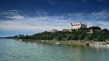 Die Burg Bratislava thront auf einem Hügel über der Donau mit Booten am Ufer und modernen Gebäuden im Hintergrund.