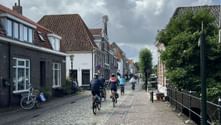 Group of cyclists riding on a cobblestone street in historic Elburg with traditional Dutch houses, red tile roofs, and green trees.