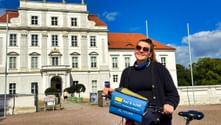 Frau mit Fahrrad und blauer Tourentasche vor dem weißen barocken Oranienburger Schloss unter blauem Himmel.