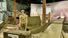 Militärausstellung mit Soldatenpuppe in Uniform neben grünem Boot mit Aufschrift 'Roer Crossing', Holzpfählen und historischen Fotos im Hintergrund.