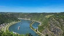 Panoramablick auf den Rhein, der sich durch steile, bewaldete Talhänge schlängelt. Frachtschiffe fahren auf dem blauen Wasser, Weinberge und Dörfer sind sichtbar.