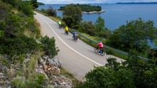 Vier Radfahrer in bunten Trikots fahren entlang einer Küstenstraße in Korčula, mit blauer Adria, kleinen Inseln und grüner Vegetation.