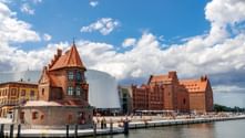 Historic brick buildings with red-tiled roofs along the harbor in Stralsund, featuring a tower and modern white structure under blue sky.