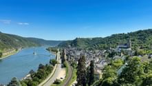 Stadt Oberwesel am Rhein mit historischen Gebäuden, Kirchtürmen, Bahnlinie am Flussufer und grünen Hügeln unter blauem Himmel.