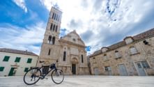 Ein Fahrrad steht auf einem Steinplatz vor der Kathedrale von Hvar mit ihrem hohen Glockenturm. Historische Steingebäude umgeben den Platz unter blauem Himmel.
