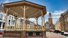 Wooden bandstand with ornate Victorian details on a brick base in Kampen's market square, surrounded by historic buildings and a church tower.