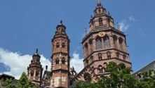Drei rote Sandsteintürme des Mainzer Doms ragen über grüne Bäume vor blauem Himmel mit weißen Wolken.