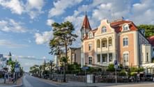 Elegant pastel-colored historic buildings with red tile roofs and a church tower line the promenade in Zinnowitz under a blue sky with clouds.