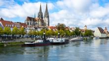 Der Regensburger Dom mit gotischen Zwillingstürmen prägt die historische Stadtsilhouette an der Donau. Ein Passagierboot fährt auf dem Wasser.