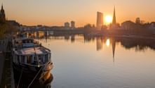 Flusskreuzfahrtschiff am Frankfurter Ufer bei Sonnenuntergang. Die Sonne spiegelt sich im ruhigen Wasser mit Skyline und Kirchtürmen.