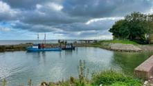 Blaues Fischerboot im ruhigen Hafenkanal von Medemblik. Grüne Grasufer säumen den Wasserweg unter dramatischem bewölktem Himmel.