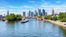 Frankfurt am Main Skyline mit modernen Wolkenkratzern und historischem roten Kirchturm, gesehen über den Main mit Frachtschiff und Steinbrücke.