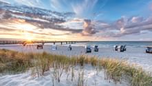 Sandy beach on Rügen with beach chairs and dune grass in foreground. A wooden pier extends into the Baltic Sea under a dramatic sunset sky.
