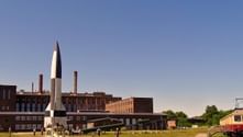 V-2 rocket replica displayed at Historical-Technical Museum Peenemünde with former power plant buildings and visitors on grass field.