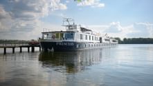 White river cruise ship MS Princess moored at wooden dock on calm water under blue sky with white clouds and green trees in background.