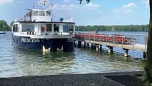 Weiß-dunkelblaues Passagierschiff MS Princess an einem Holzsteg mit roten Geländern auf einem See, umgeben von grünen Bäumen unter bewölktem Himmel.