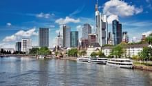 Frankfurt am Main skyline with modern skyscrapers and historic buildings along the Main River. Tour boats are docked at the waterfront under blue sky.