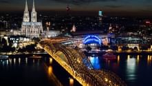 Nachtansicht von Köln mit beleuchteter gotischer Kathedrale und Hohenzollernbrücke über den Rhein, Stadtlichter spiegeln sich im Wasser.