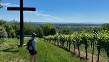 Wanderer mit Rucksack steht bei Holzkreuz auf grasigem Hang mit Weinreben und Panoramablick ins Tal unter blauem Himmel.