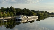 White Princess river cruise ship moored at a wooden dock in Schwedt. The vessel is reflected in the calm water, surrounded by green trees under a cloudy sky.