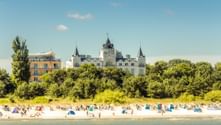 Sandy beach in Zinnowitz with colorful beach chairs and visitors. White castle-like building with towers behind green trees under blue sky.