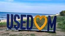 Large blue and orange USEDOM letters with heart symbol on sandy beach. Baltic Sea and dune grass visible in background under blue sky.