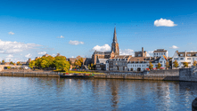 Maastricht cityscape with historic church spire and colorful buildings along the Maas River under a blue sky with white clouds.