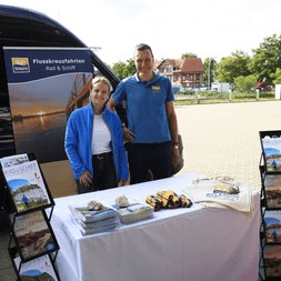 Two staff members in blue shirts at an outdoor booth with brochures and promotional materials for river cruises on a white-covered table.