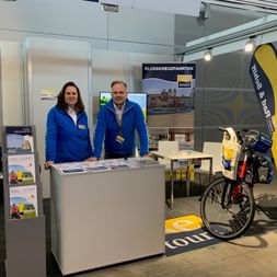 Zwei SE-Tours Mitarbeiter in blauen Jacken an ihrem Messestand mit Broschüren, Displays und einem Reiserad mit Packtaschen.