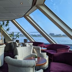 Interior of MS SE-MANON lounge featuring gray and purple seating with round tables, large angled windows offering water views, and green plants.