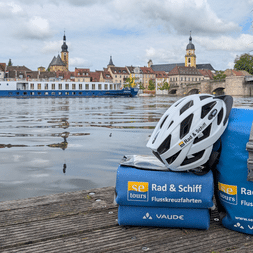 Blue Rad & Schiff cycling bags and white helmet on wooden dock. Historic town with church towers and Casanova ship visible across the Danube.