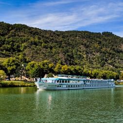 Weißes Flusskreuzfahrtschiff MS Switzerland fährt auf ruhigem grünen Wasser mit bewaldeten Hügeln und Dorfgebäuden im Hintergrund unter blauem Himmel.
