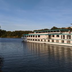 White river cruise ship Switzerland with green trim sailing on calm river. Trees line the left bank, buildings visible on distant hills under blue sky.