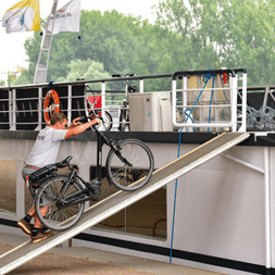 Person schiebt ein Fahrrad eine Metallrampe hinauf auf das Nassau-Schiff. Das weiße Schiff hat Geländer und Fenster, Flaggen im Hintergrund sichtbar.