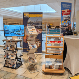 Trade fair booth with Kempf branding, brochure displays, and promotional materials in a shopping center. A monitor and banners advertise river cruises.