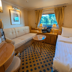 Bright cabin with beige sofa, single bed, wooden furniture, patterned carpet, and window with gold curtains showing river view.