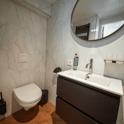 Kompaktes Badezimmer auf Magnifique X mit weißen Marmorwänden, Wandtoilette, dunklem Waschtisch mit Waschbecken, rundem Spiegel und Holzboden.